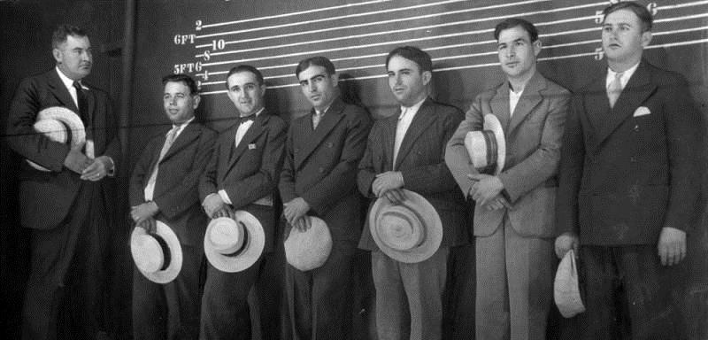 Photo shows six asserted racketeers lined up in the Los Angeles police shadowbox today while detectives look them over in effort to link them with six gang murders. The men stand behind a thin screen in a stage-like box. It is illuminated so the suspects can be clearly seen by detectives, but cannot see the detectives inspecting them from the darkness. Left to right, Detective Lieutenant ‘Lefty’ James, John Testini, Dominic de Soto (aka Domenico Di Ciolla), alleged rival of slain August Palombo; Vito Cisareo, Domenic Shiovone, Salvadore Verna, Louis Tasselli.” Los Angeles Herald Examiner, July 26, 1928.